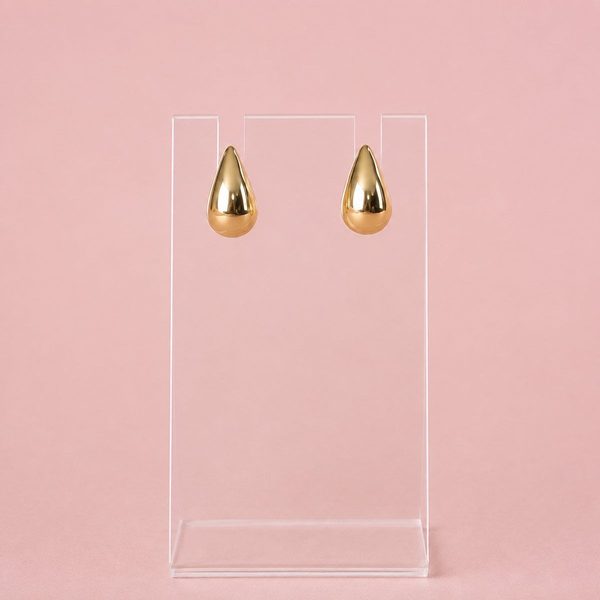 A pair of gold teardrop-shaped drop earrings displayed on a clear acrylic stand against a solid pink background.