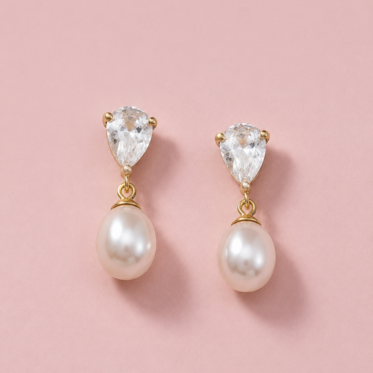 A pair of gold drop earrings featuring pear-cut clear crystals at the top and smooth white freshwater pearls hanging below, displayed on a soft pink background.