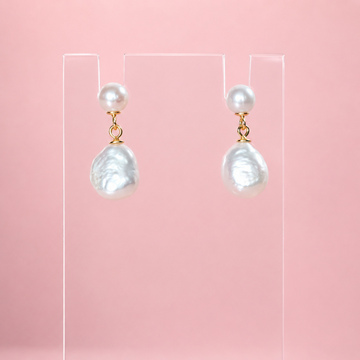 A pair of baroque pearl drop earrings with gold, displayed on a clear acrylic stand against a solid pink background.