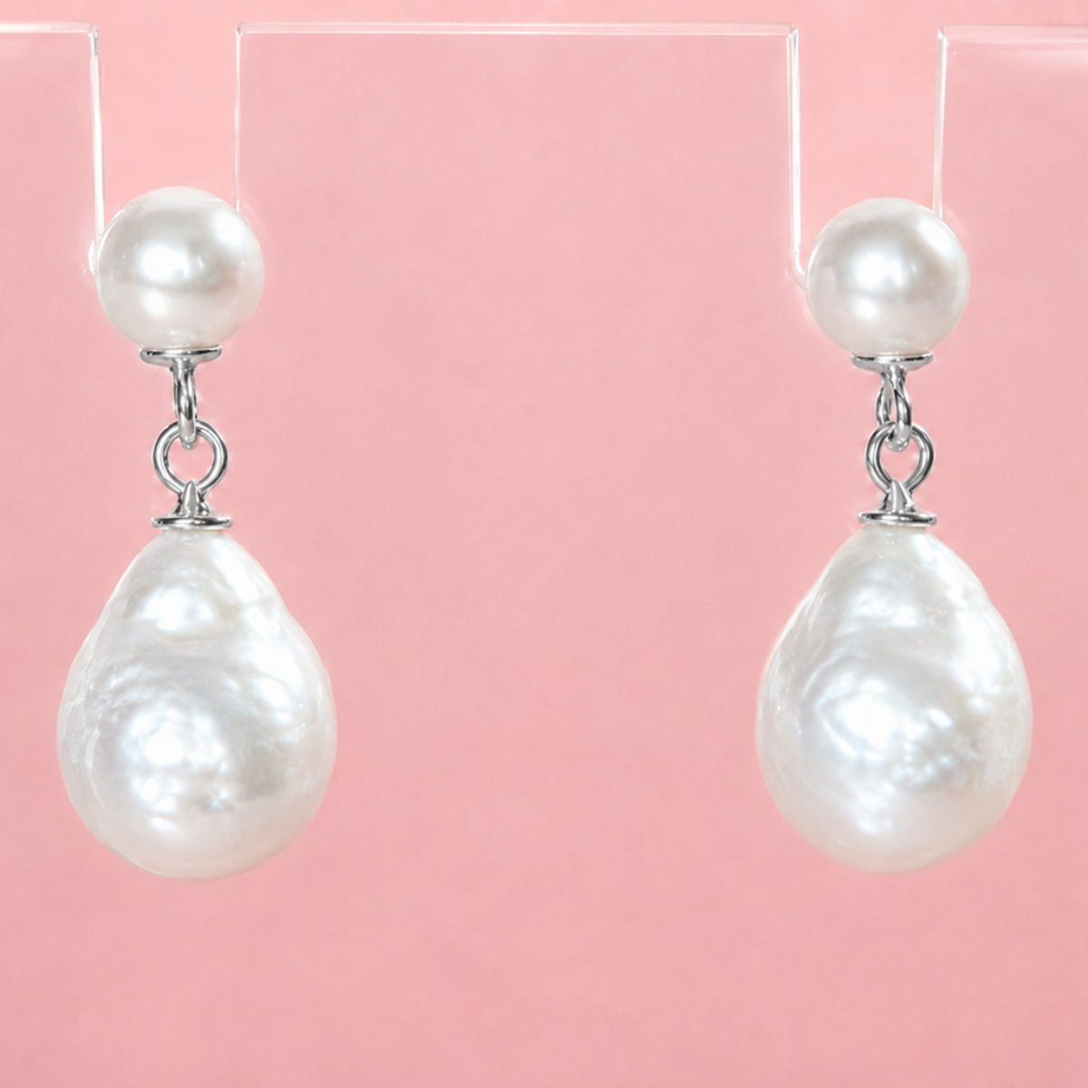 A pair of white freshwater pearl drop earrings with teardrop-shaped pearls and silver-toned hooks, displayed on a clear acrylic stand against a solid pink background.