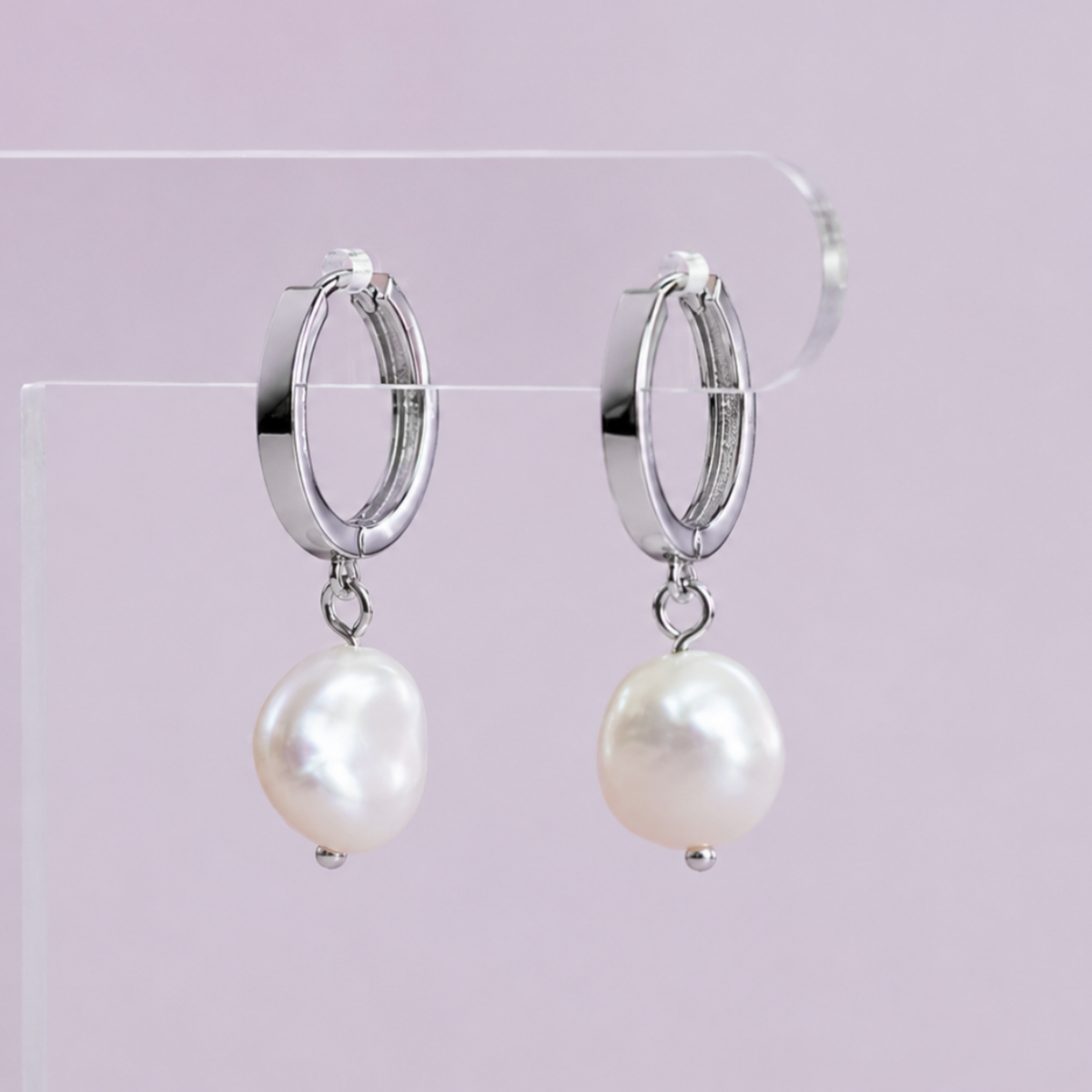A pair of silver hoop earrings with dangling baroque-style white pearls, displayed on a clear acrylic stand against a soft pink background.