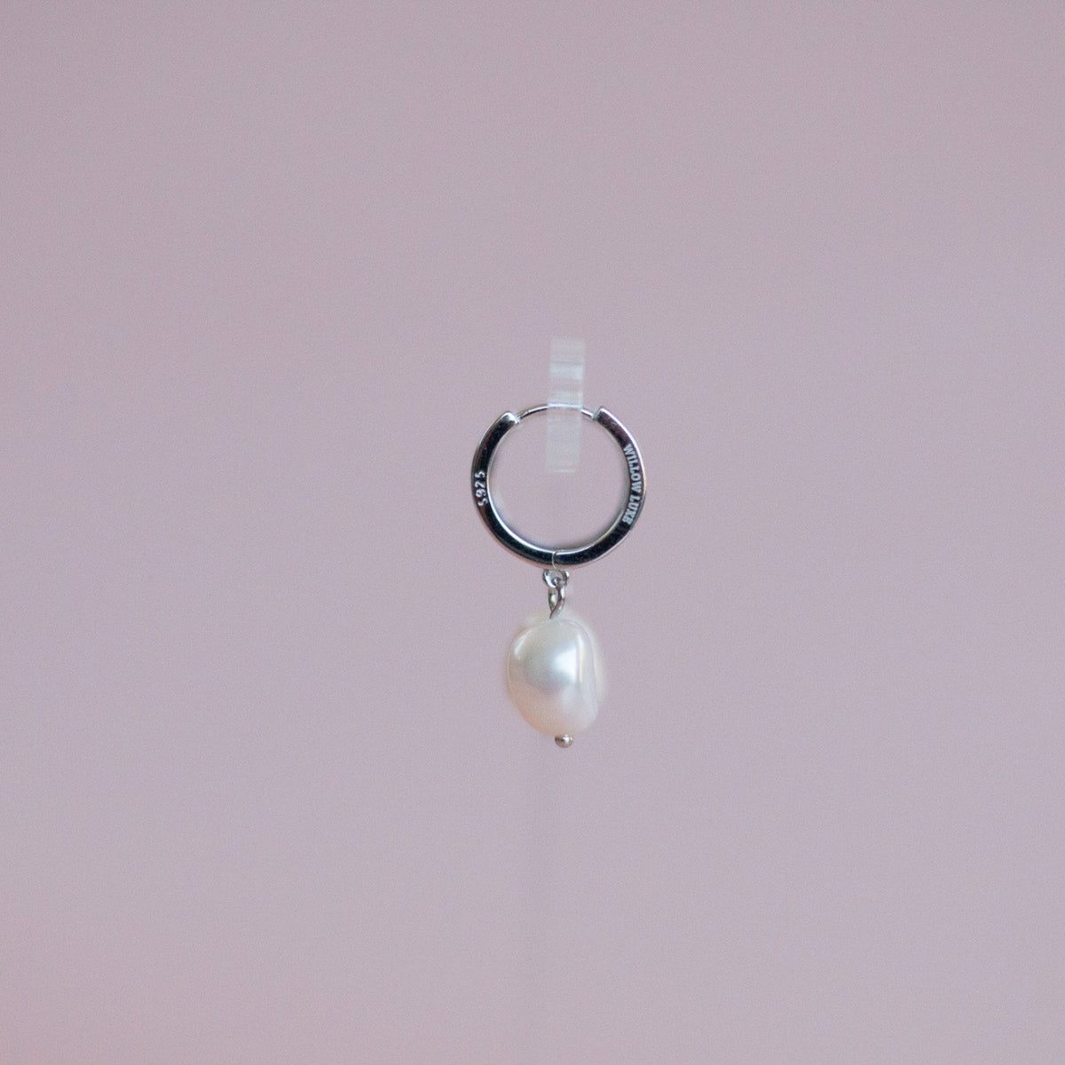 A silver huggie hoop earring with a dangling baroque pearl, suspended against a solid light pink background.
