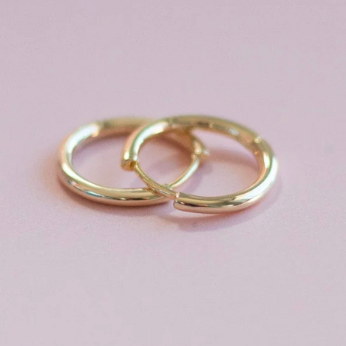 Two small gold hoop earrings overlapping on a soft pink background.