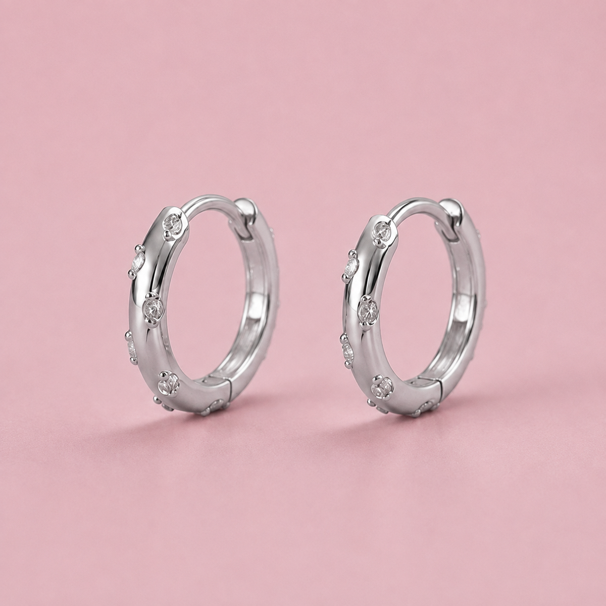 A pair of polished silver hoop earrings with small clear gemstones set along the front, displayed on a solid pink background.
