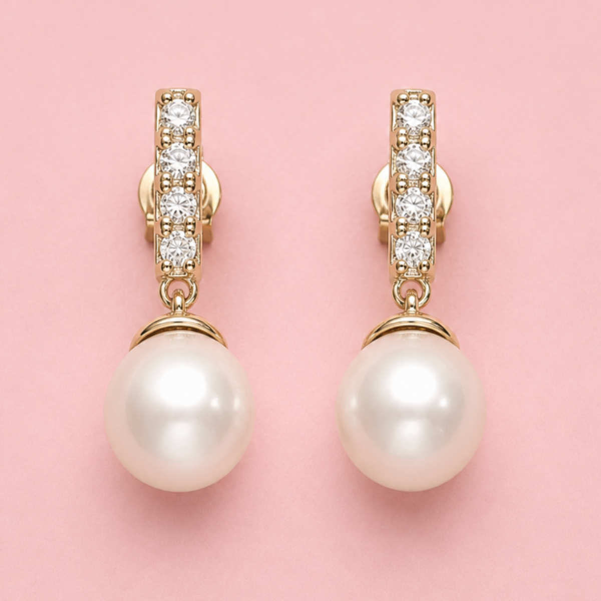 A pair of gold drop earrings with a vertical bar set with clear stones and a large white pearl dangling below, shown on a solid pink background.