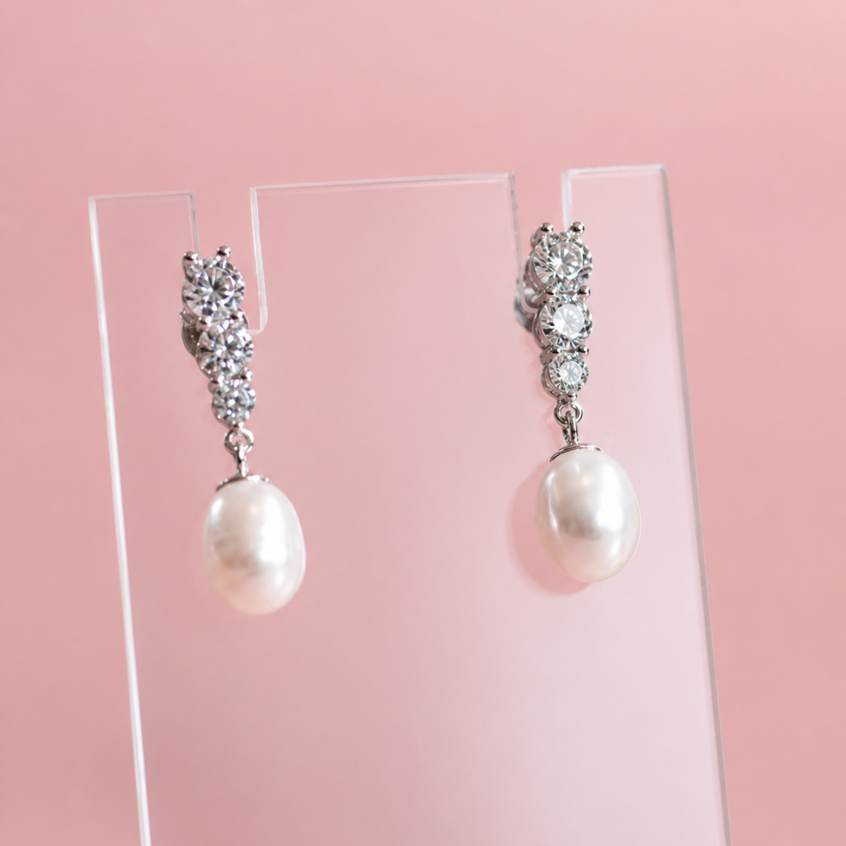A pair of silver-tone drop earrings with clear crystal clusters at the top and a single oval white pearl hanging below, displayed on a clear acrylic stand against a soft pink background.