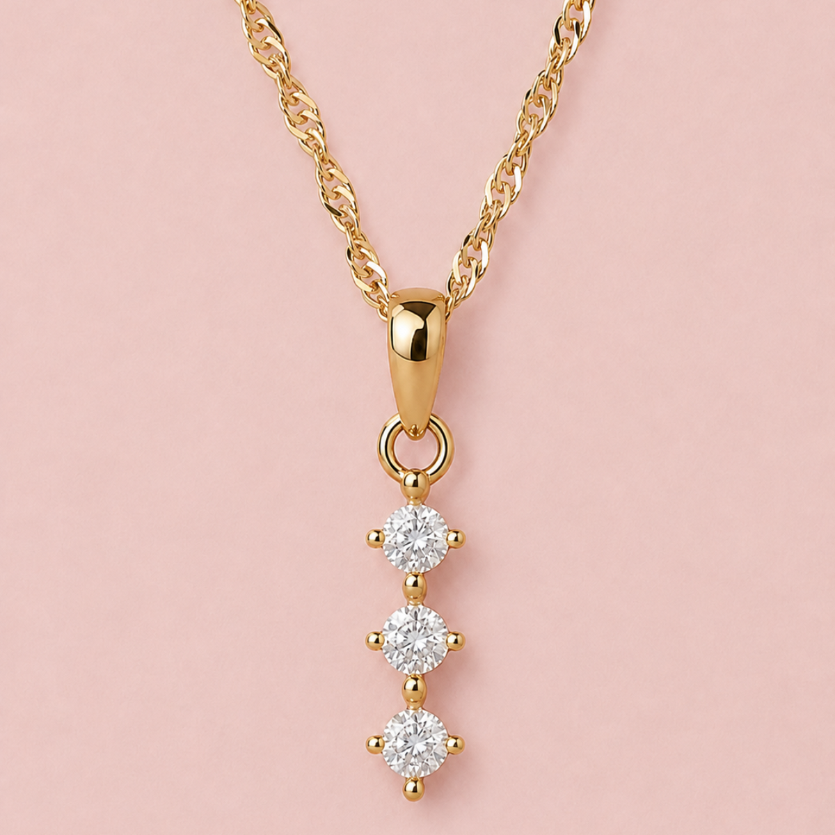 A gold necklace with a vertical dangle pendant featuring three round-cut clear gemstones in a prong setting, displayed against a solid pale pink background.