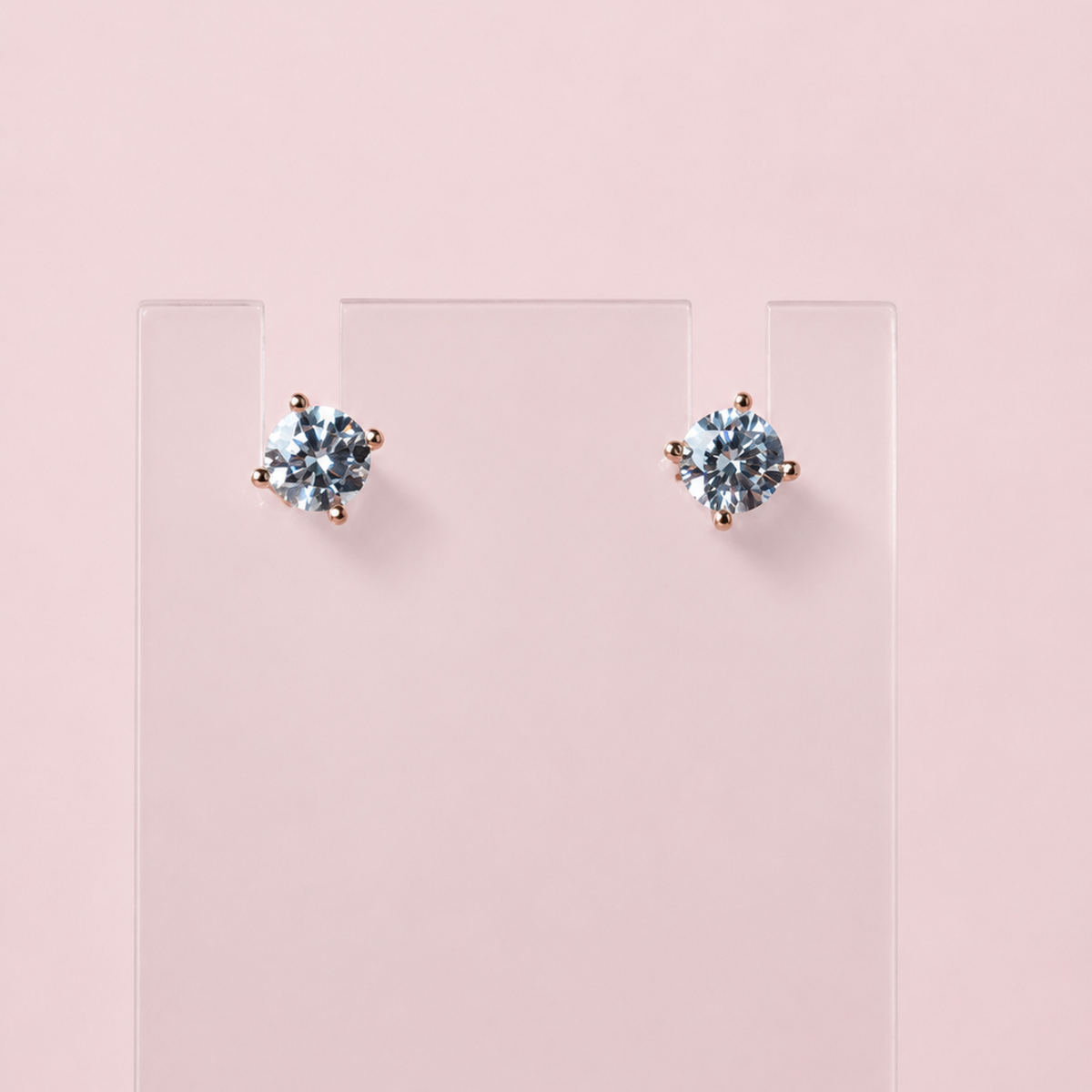 A pair of stud earrings with round light blue gemstones in a prong setting, displayed on a clear rectangular stand against a soft pink background.