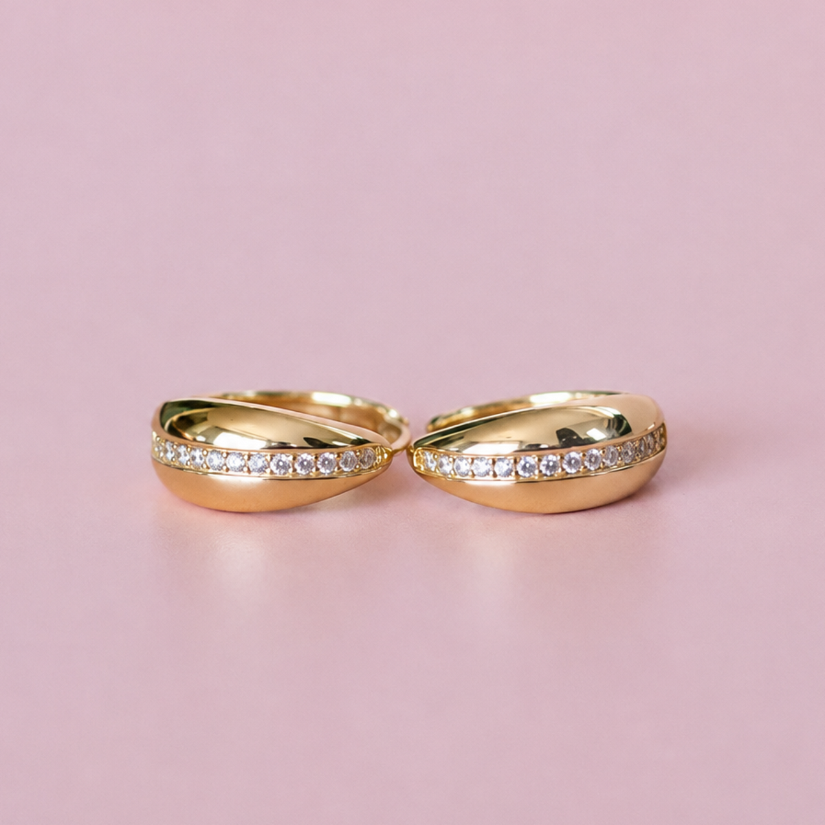 Two gold hoop-style rings with rows of small clear stones, displayed on a soft pink background.
