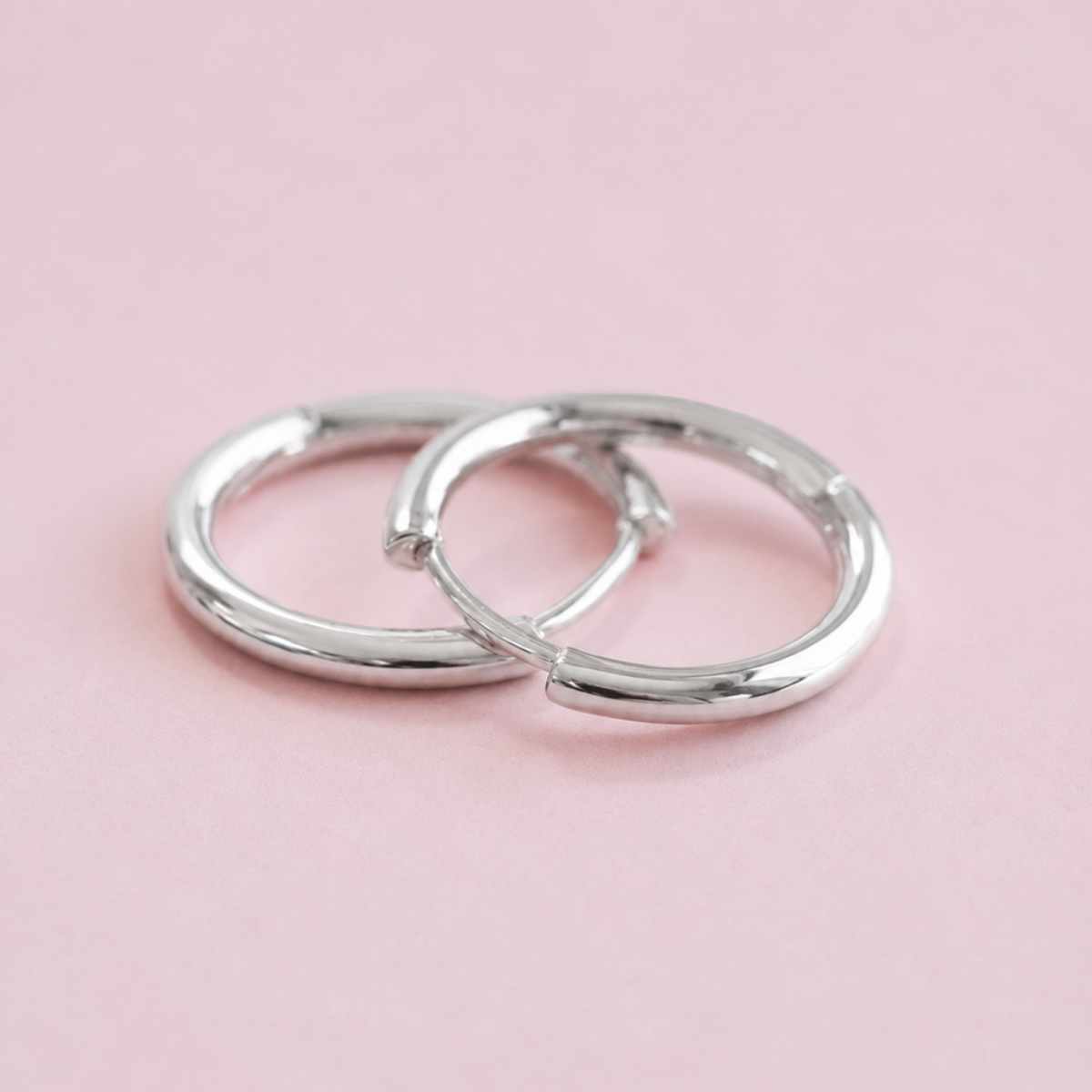 A pair of polished silver hoop earrings resting on a solid light pink background.