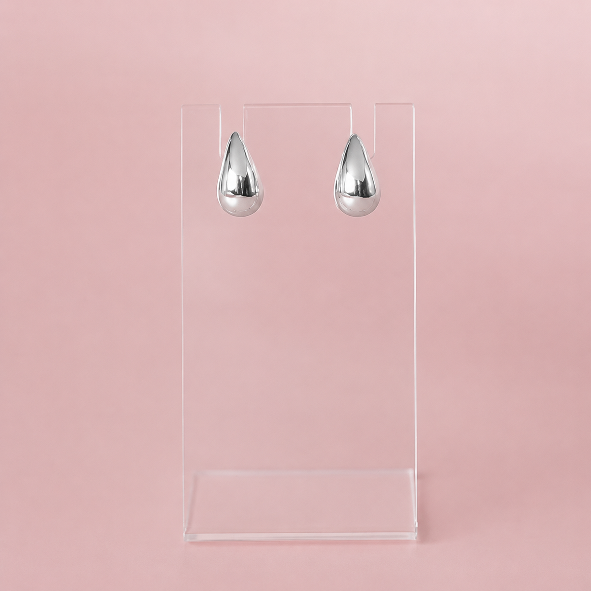 A pair of shiny silver teardrop-shaped earrings displayed on a clear acrylic stand against a solid pink background.