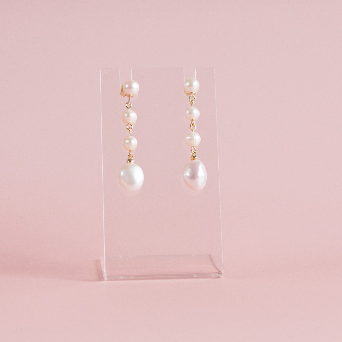 A pair of dangle earrings with white pearls and gold-tone metal, displayed on a clear acrylic stand against a soft pink background.