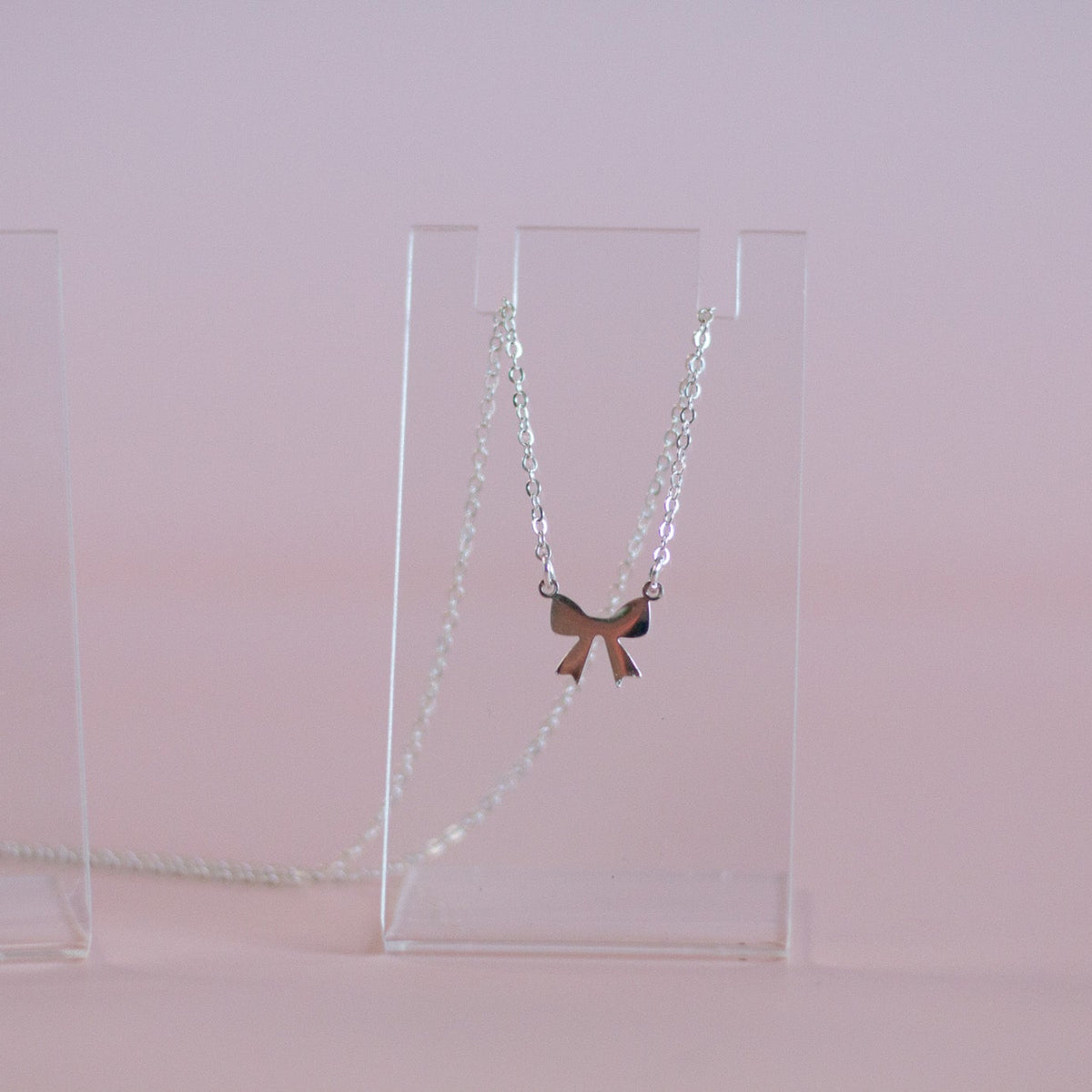 A delicate silver chain necklace with a small bow-shaped pendant, displayed on clear acrylic stands against a soft pink background.