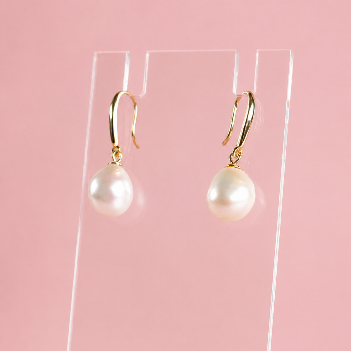 A pair of gold-tone French hook earrings with large white pearl drops, displayed on a clear acrylic stand against a solid pink background.