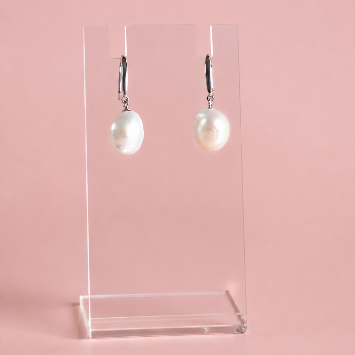 A pair of silver drop earrings with large, lustrous white baroque pearls, displayed on a clear acrylic stand against a solid pink background.