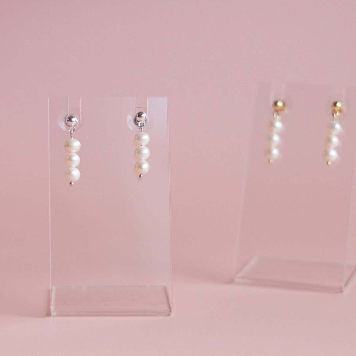 Dangle earrings with three white pearl-like beads and a small gold bead at the top, displayed on clear acrylic standins against a soft pink background.