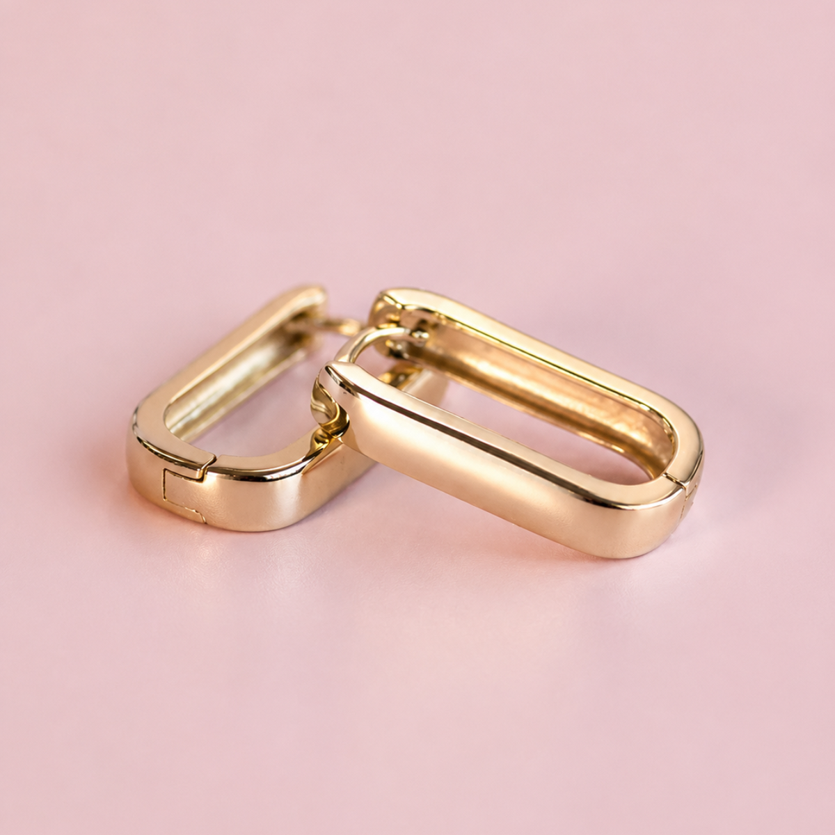 A pair of gold-toned metal clip-on earrings with a polished, rounded rectangular design, resting on a solid light pink background.