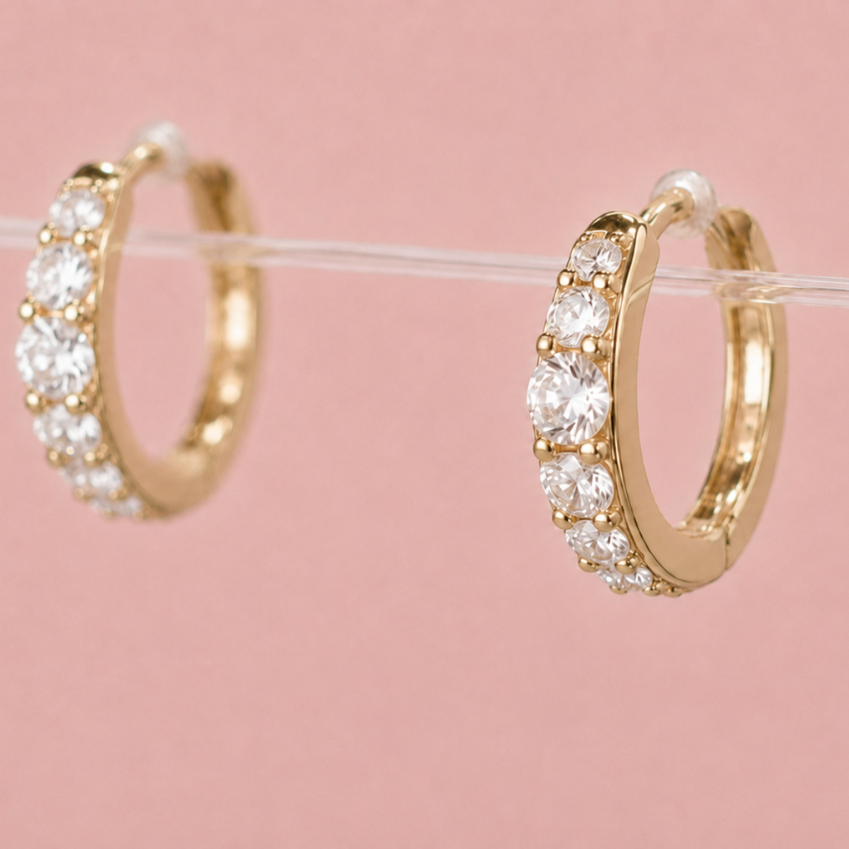 Gold hoop earrings with clear pavé-set stones, displayed on a clear stand against a soft pink background.