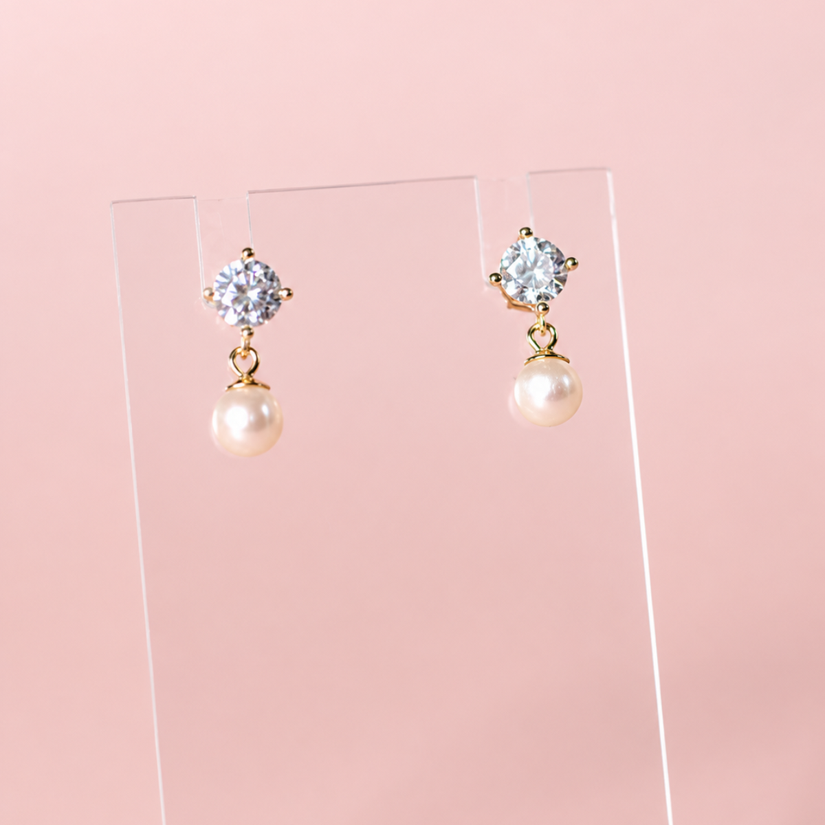 A pair of gold drop earrings with clear round-cut stones at the top and white pearls hanging below, displayed on a clear acrylic stand against a soft pink background.