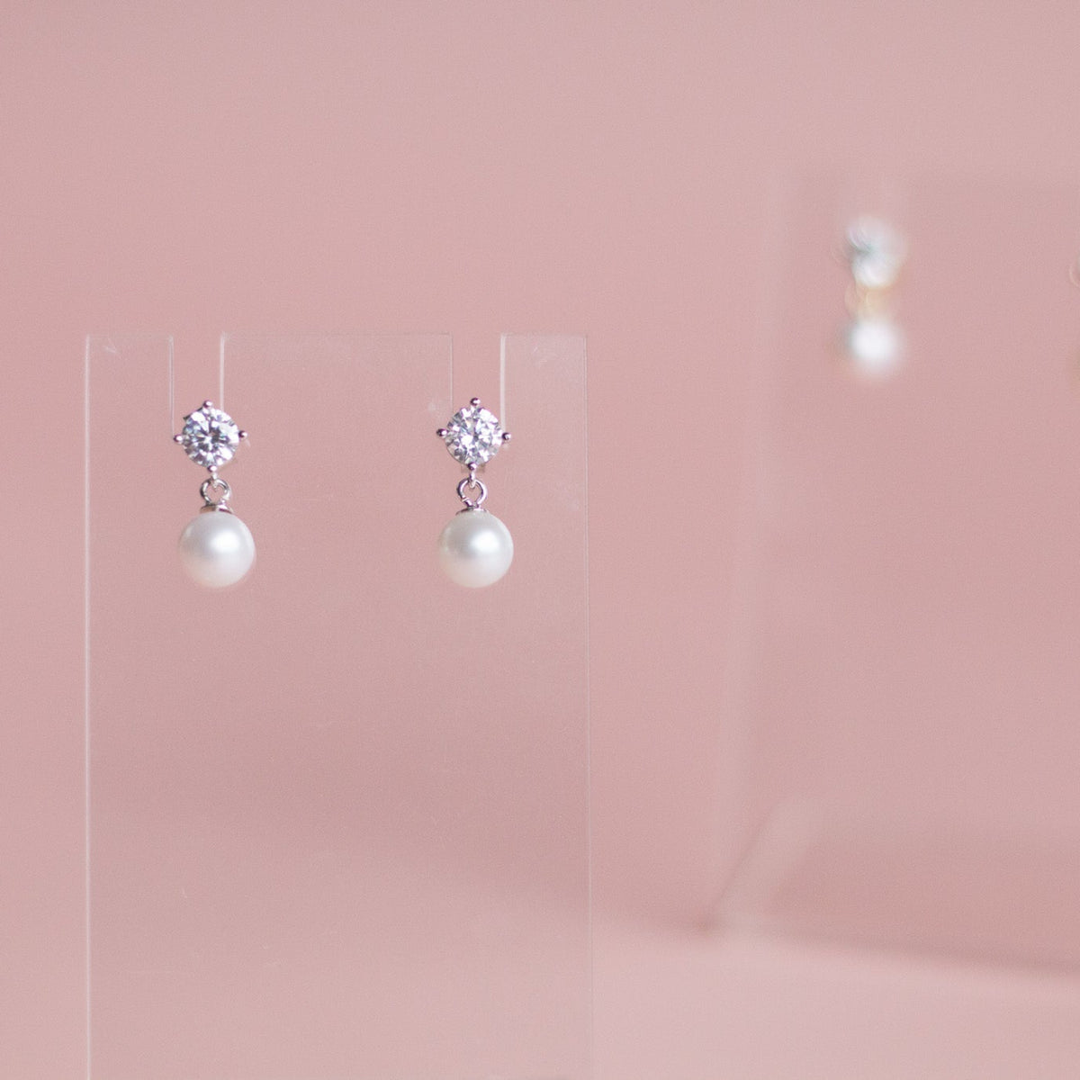 A pair of silver-toned dangle earrings with clear round-cut stones at the top and a white pearl drop below, displayed on a clear acrylic stand against a soft pink background.