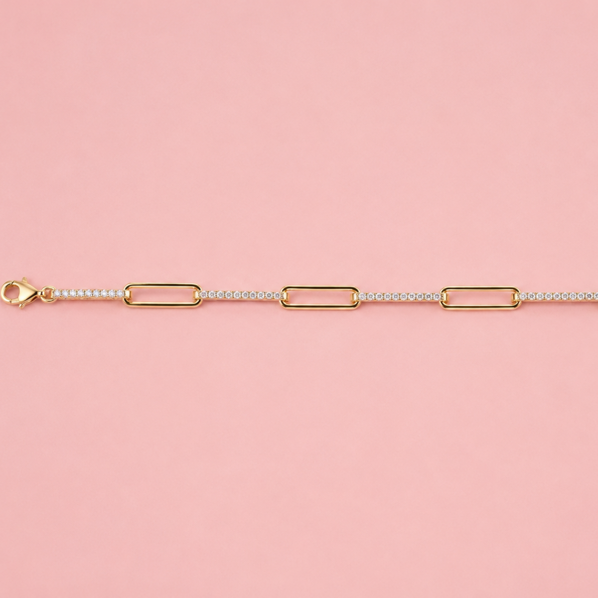 A gold chain bracelet with alternating elongated rectangular links and small round links, laid horizontally on a solid pink background.
