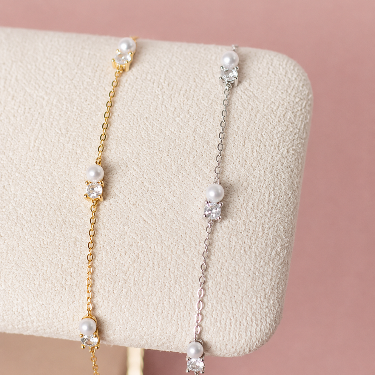 Two delicate chain bracelets displayed on a cream-colored jewelry stand against a soft pink background. One bracelet is gold-toned with white pearls and clear moissanite accents, and the other is silver-toned with matching white pearls and clear stone accents.