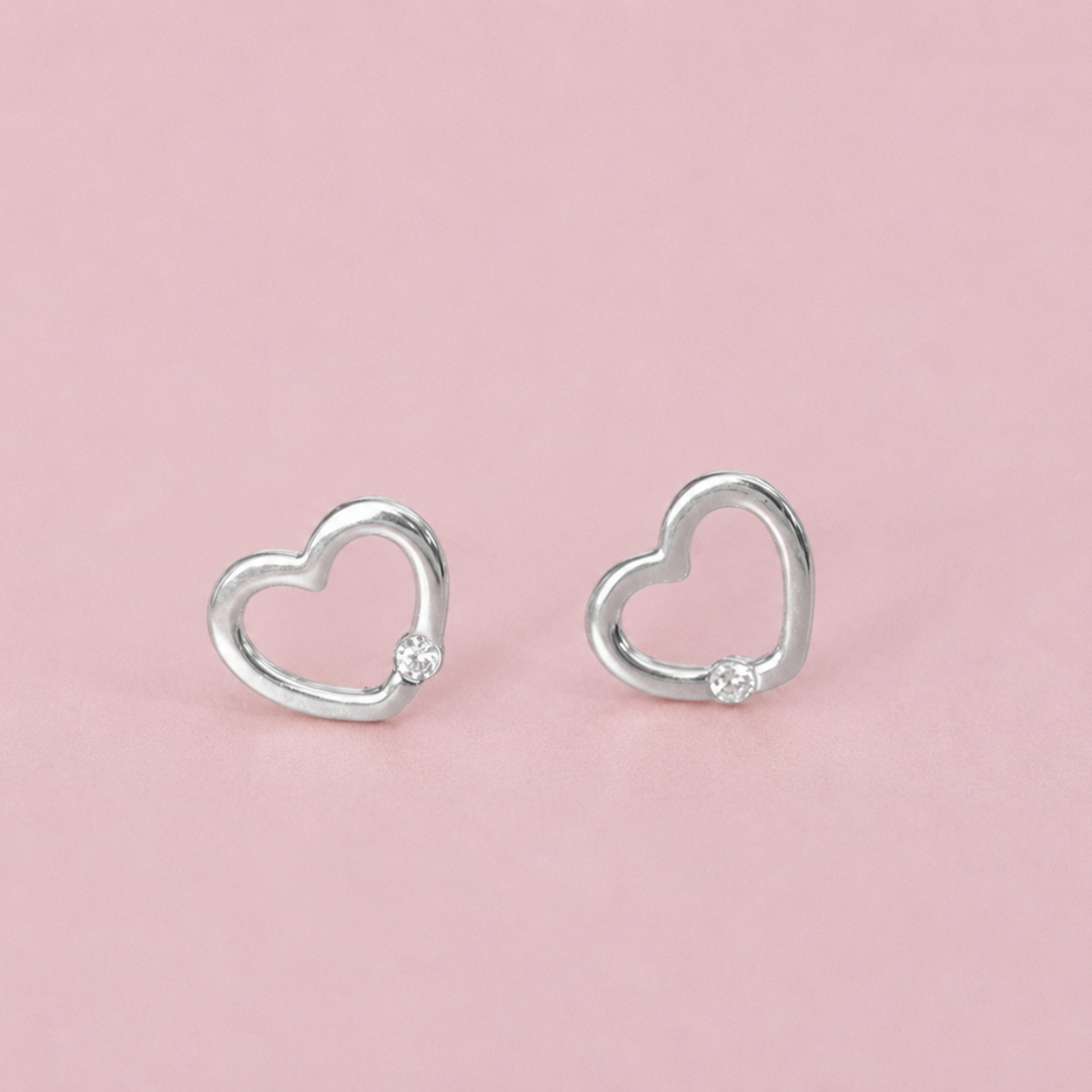 A pair of silver heart-shaped hoop earrings with a small clear gemstone at the bottom curve, displayed on a solid pink background.