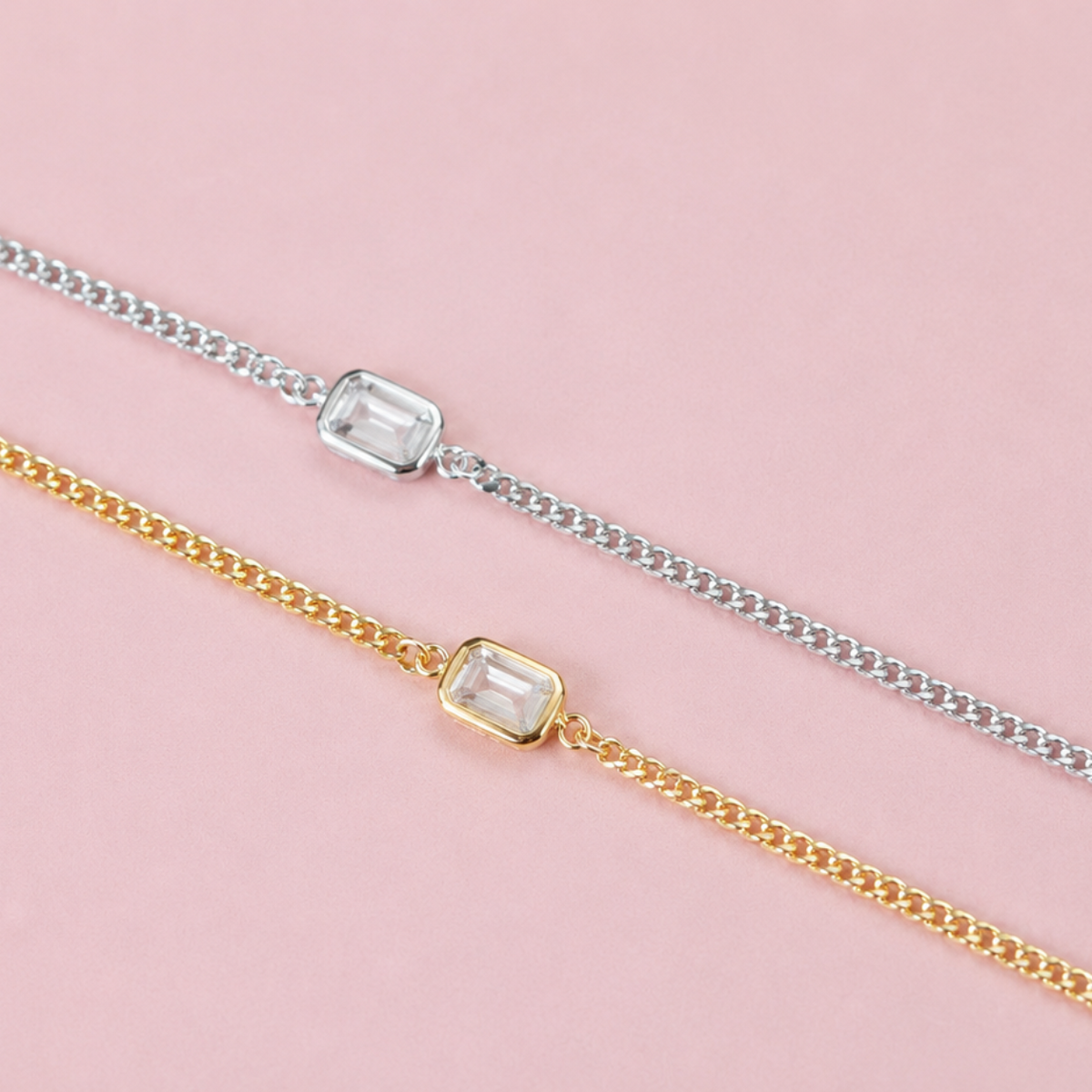 Two delicate chain bracelets, one in silver and one in yellow gold, each featuring a rectangular clear gemstone bezel setting, displayed diagonally on a solid pink background.