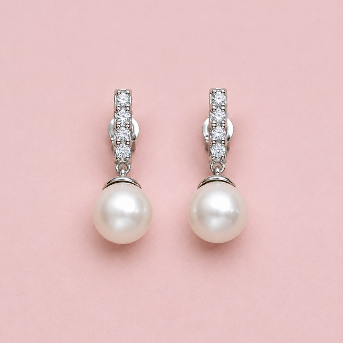 A pair of silver drop earrings with round white pearls and small clear stones in the upper section, displayed on a solid pink background.