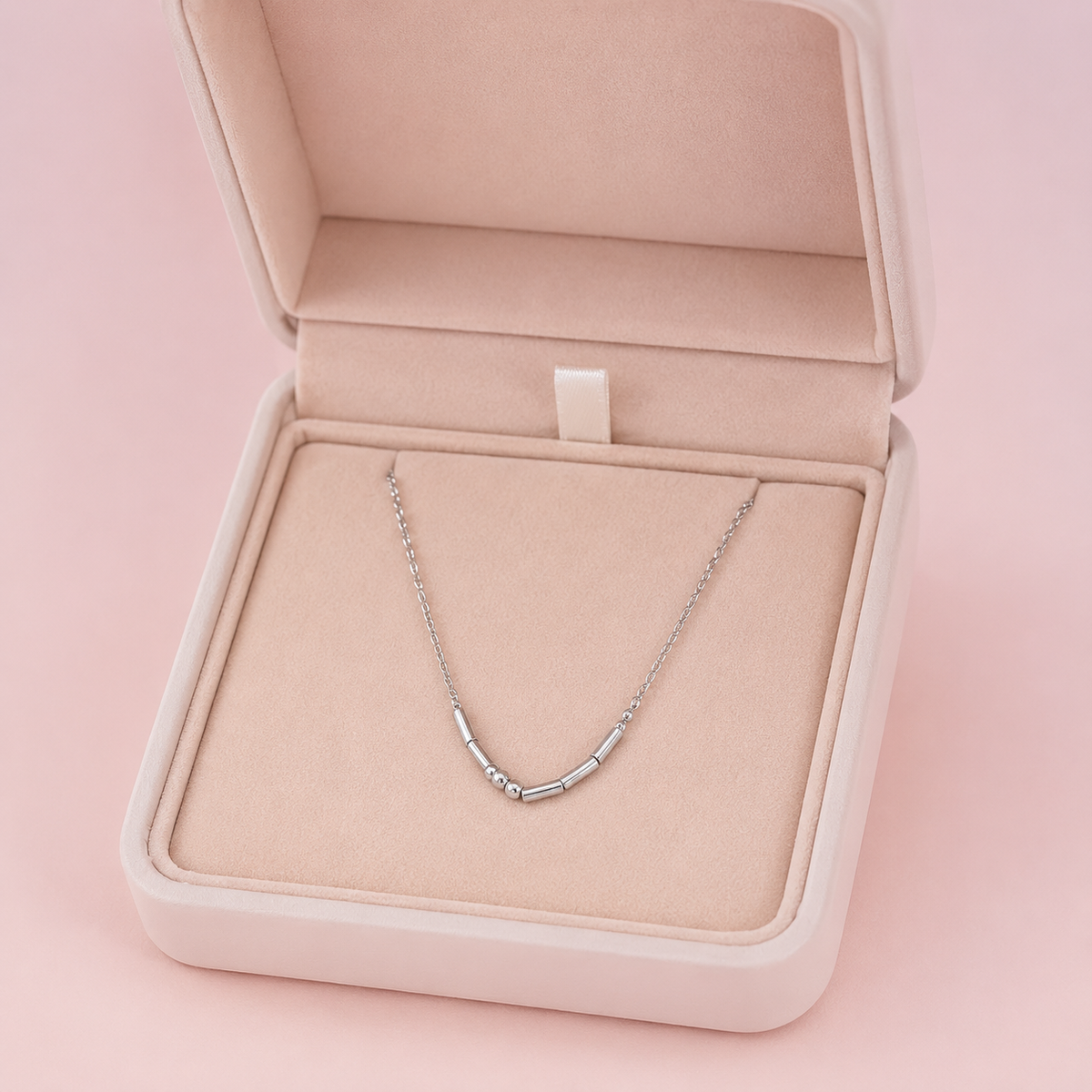 A delicate silver necklace with a curved, segmented tube design displayed inside an open blush pink jewelry box on a soft pink background.